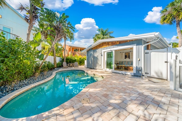 A swimming pool with a brick patio, surrounded by lush greenery and a modern house with a glass door.