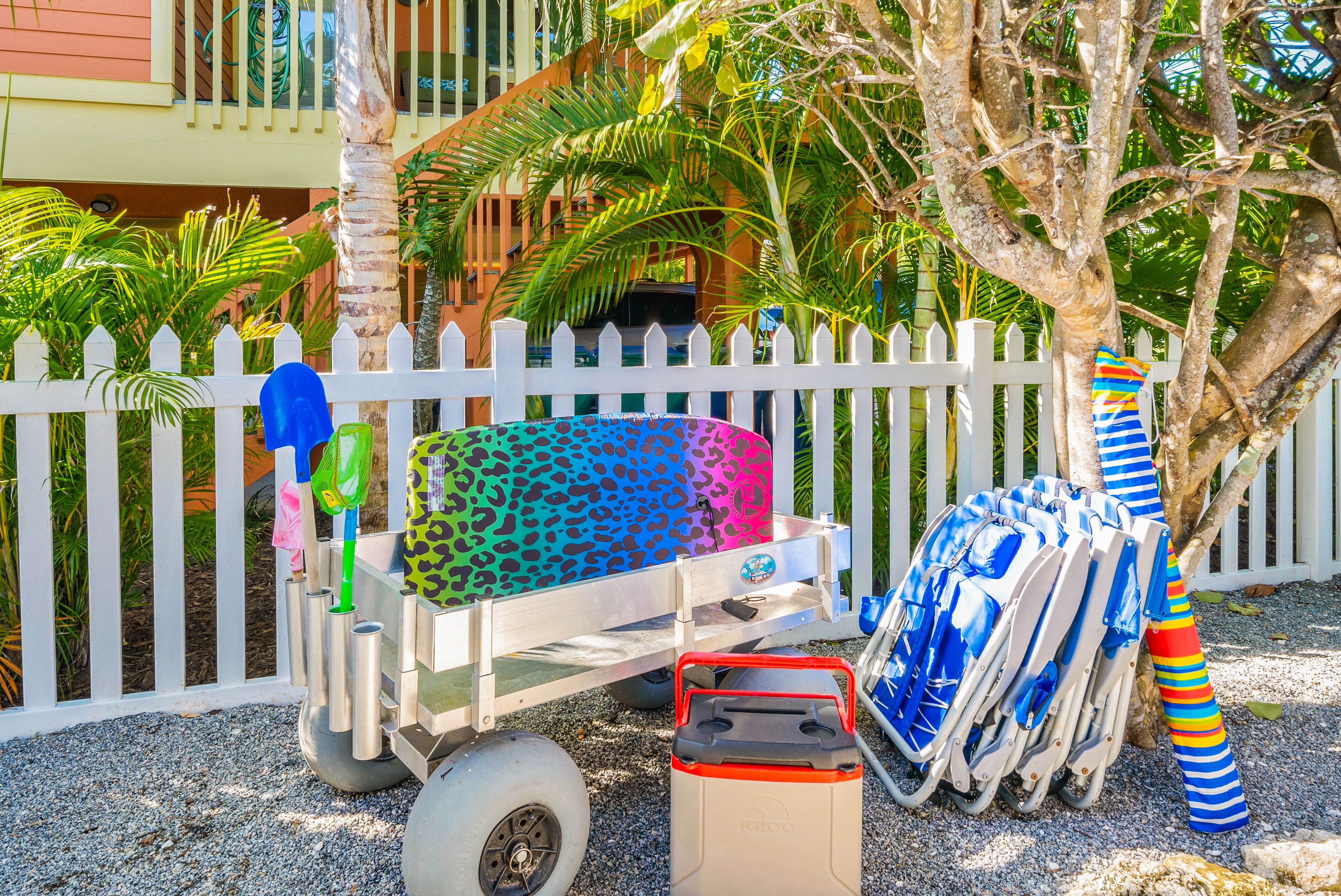 A beachside setting with a white picket fence, a beach cart with colorful beach chairs, and a red cooler.