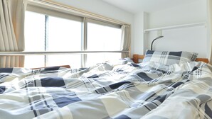 A neatly made bed with a plaid bedspread, a window with curtains, and a bedside table with a lamp. - Merveille Ueno House Tokyo (Tokyo)