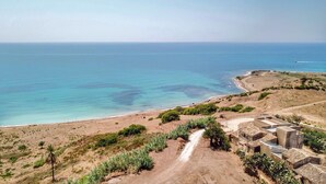 Paesaggio costiero con un sentiero sterrato che conduce a un gruppo di edifici a tetti piatti, affacciato su una spiaggia e il mare.