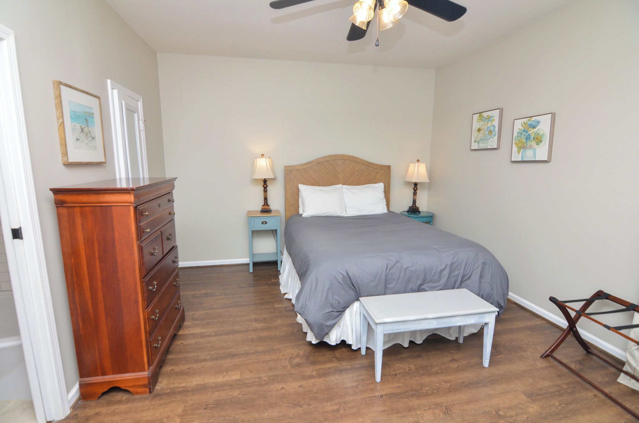 Une chambre à coucher avec une commode en bois, un lit recouvert d’une couverture grise, deux lampes de chevet et un ventilateur de plafond.
