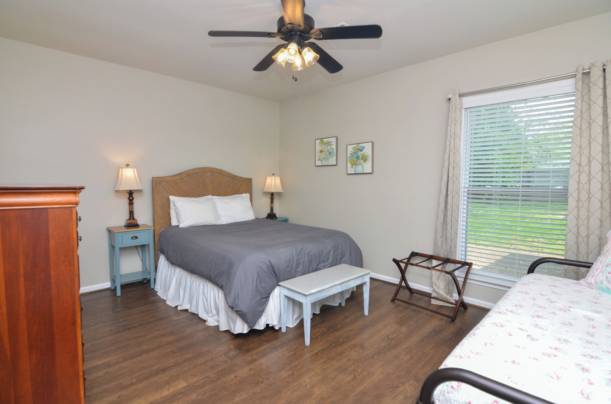 A bedroom with a bed, bedside tables, a ceiling fan, a window with blinds, and a wooden floor.
