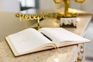 Um livro aberto sobre uma superfície de mármore, com uma caneta, um letreiro dourado com a palavra 'Reception' e uma pequena moldura de foto.