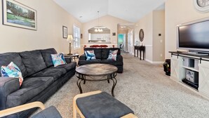 A spacious living room with a large TV, two sofas with decorative pillows, a coffee table, and a kitchen in the background.
