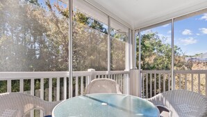 A covered outdoor area with wicker chairs and a glass table, surrounded by white railings and trees.