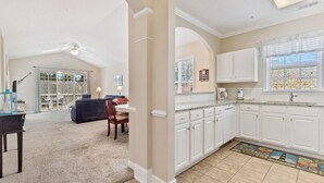 A spacious living room with a dark sofa, a dining area with a table and chairs, and a kitchen with white cabinets and a window with blinds.