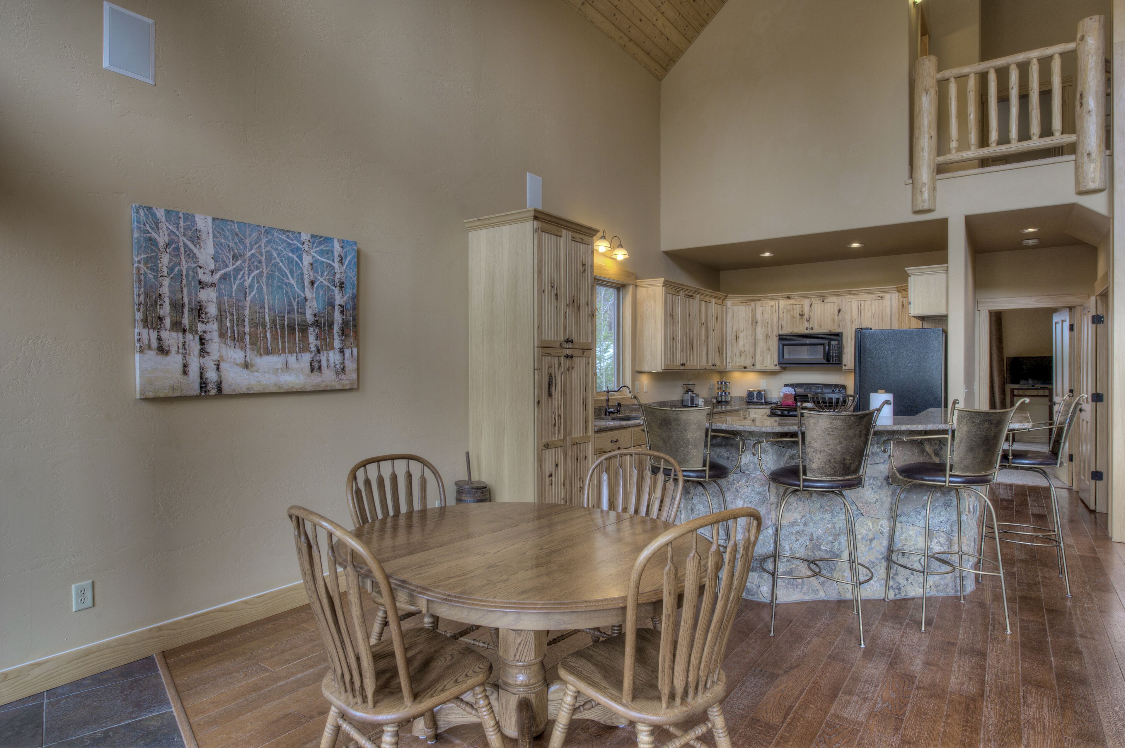 A spacious kitchen with wooden cabinets, a dining area with a large wooden table and chairs, and a painting of a snowy forest on the wall.