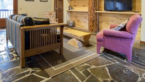 Ein gemütliches Wohnzimmer mit Steinboden, Holzbalkendecke, einem an einer Holzwand befestigten Fernseher, einer hölzernen Bank und einem violetten Sessel.