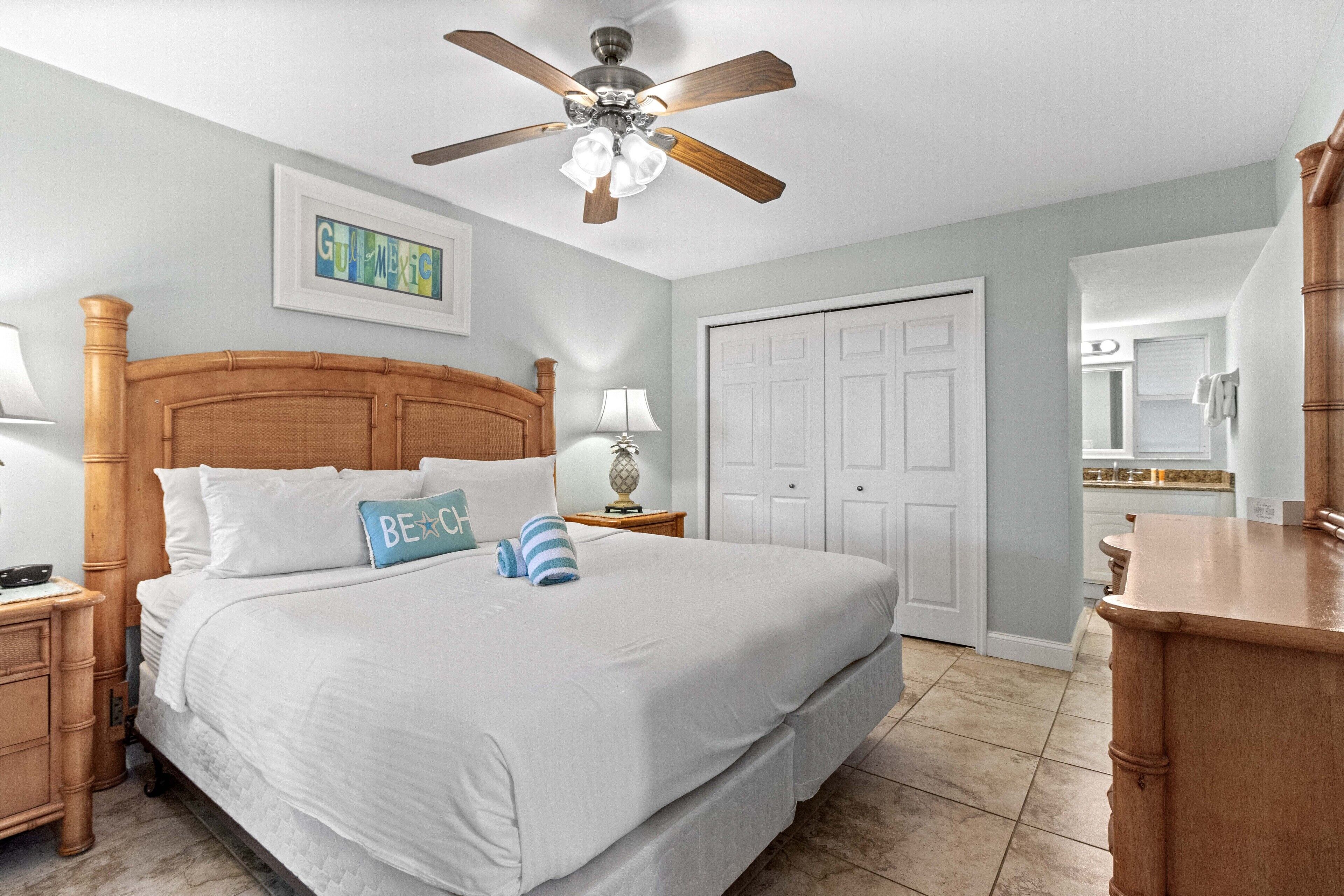 A hotel room with a wooden headboard, a bed with white bedding, a ceiling fan, a framed artwork on the wall, and a white door.