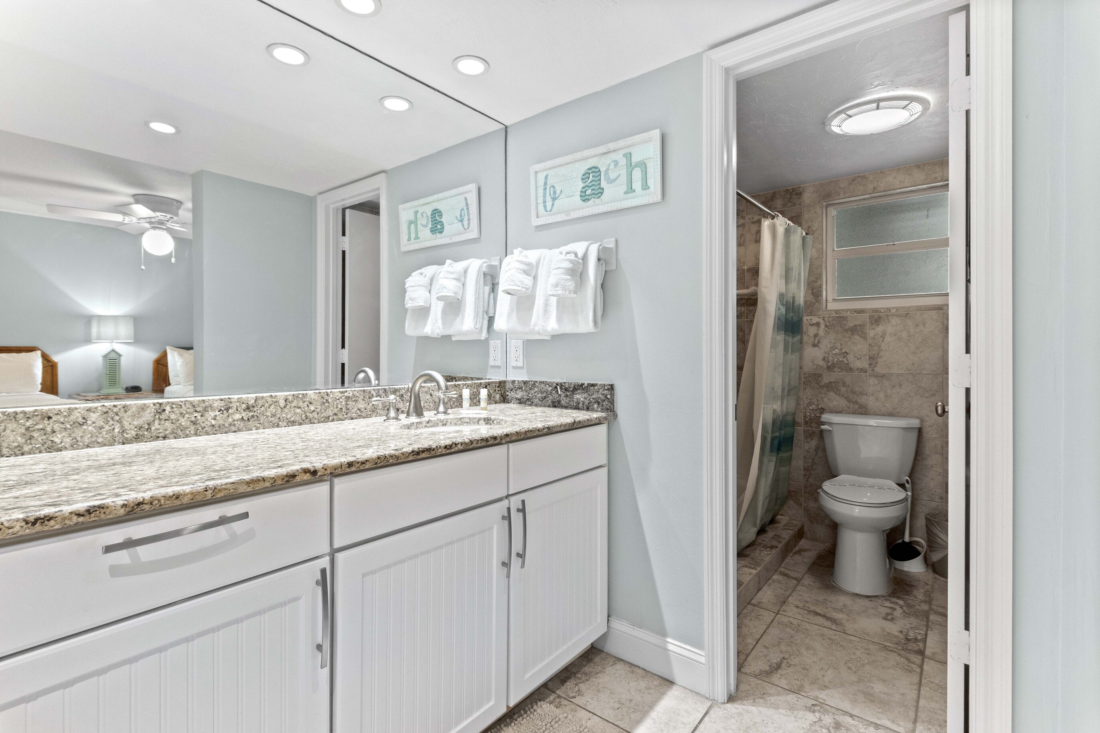 A modern bathroom with a granite countertop, white cabinets, and a large mirror. There are two towel racks with white towels, a shower area with a glass door, and a toilet.