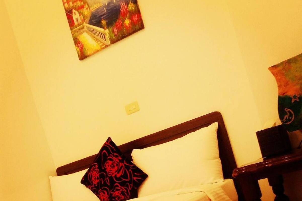 A bed with a white sheet and a black rose-patterned pillow, a wooden headboard, and a painting on the wall.