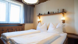 Una cama doble con sábanas blancas, cabecera de madera y dos mesitas de noche. Se ve una ventana con cortinas, una estantería con adornos y lámparas fijadas a la pared.