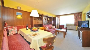 Una sala de estar acogedora con un sofá, una mesa con una botella y fruta, un televisor y una ventana con cortinas.