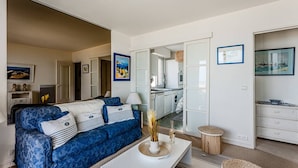 A modern living room with a blue sofa, a white coffee table, and a kitchen area visible through an open door.