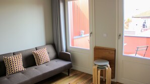 Ein Zimmer mit einem grauen Sofa, gestreiften Kissen, einem Holzschemel und einem Fenster mit Blick auf ein rotes Gebäude.