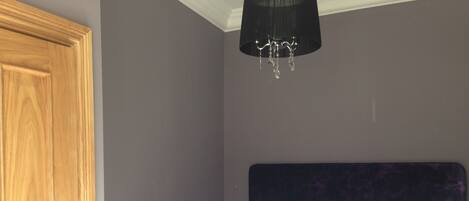 Un lit bien fait, avec du linge de lit blanc et deux coussins violets, une porte en bois et un lustre.