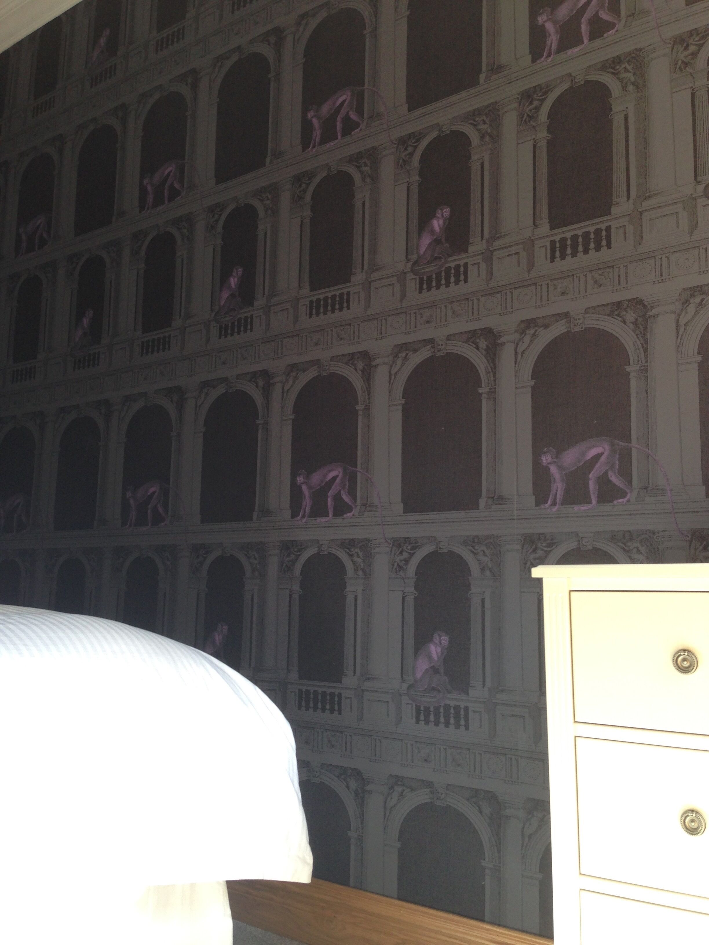 A room with a large mural of classical architecture and statues, a white bed, and a wooden dresser.