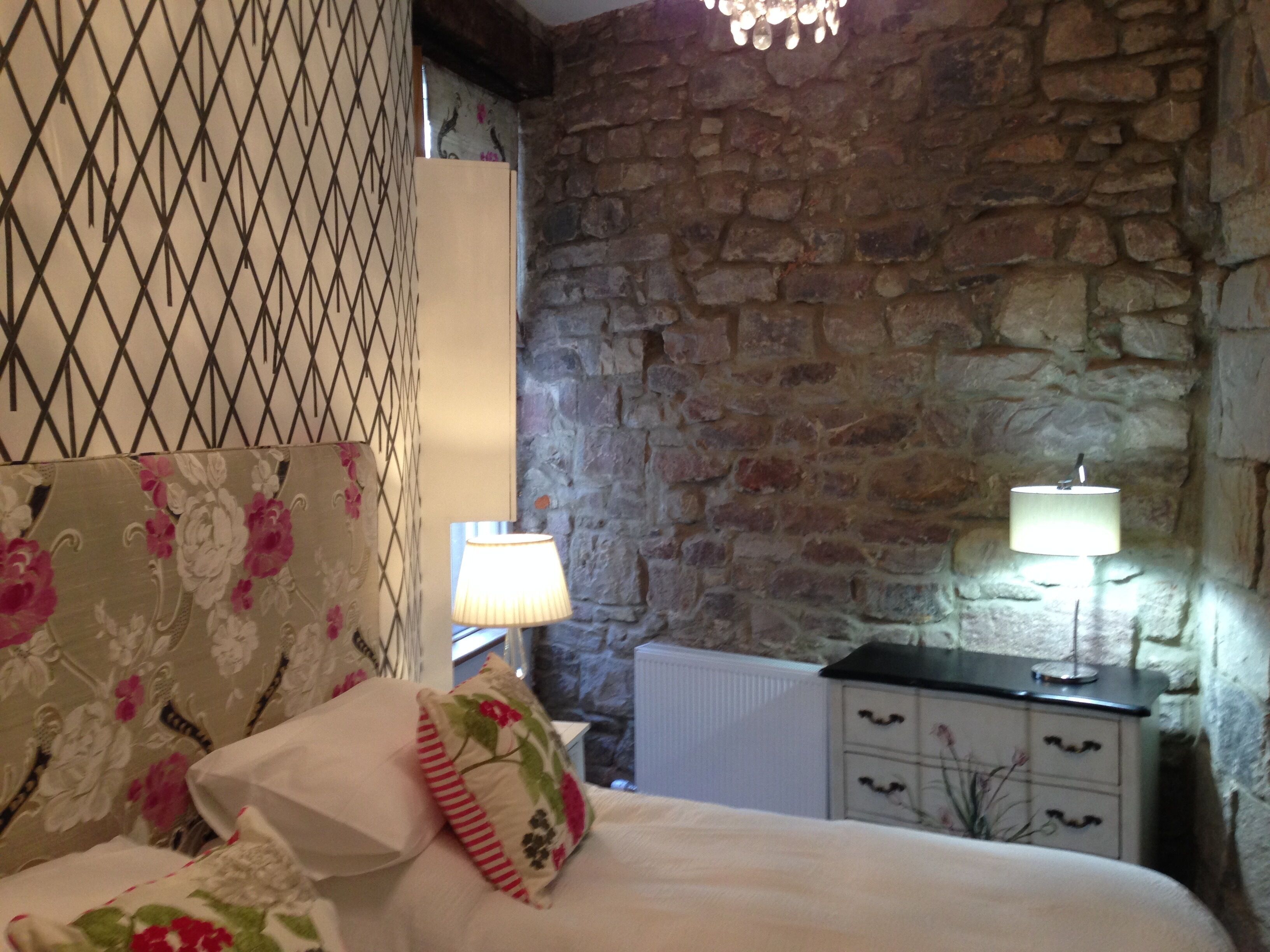A bedroom with a stone wall, a bed with floral bedding, two bedside lamps, and a radiator.