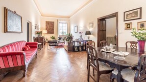 Un salon spacieux avec un canapé rouge, une table à manger en bois et des chaises. On y trouve une cheminée, un coin repas avec une table dressée, et une grande fenêtre.