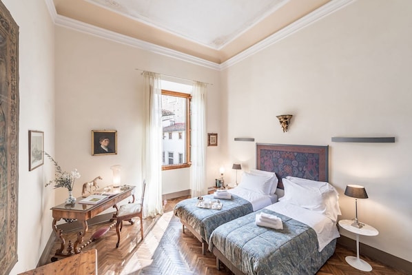 A hotel room with two beds, a desk with a lamp, a chair, a window with curtains, and a painting on the wall.