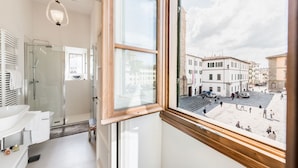 Une salle de bain avec une douche en verre, un lavabo et une fenêtre donnant sur une rue bordée d’immeubles.