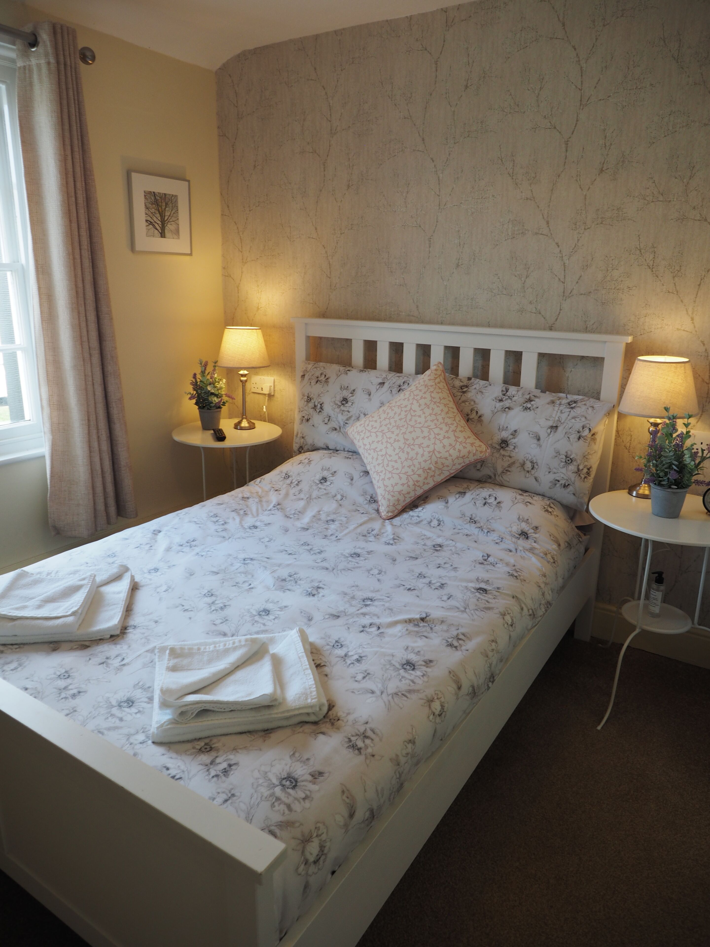 A neatly made bed with floral bedding, two bedside tables with lamps, a potted plant on each table, and a framed picture on the wall.