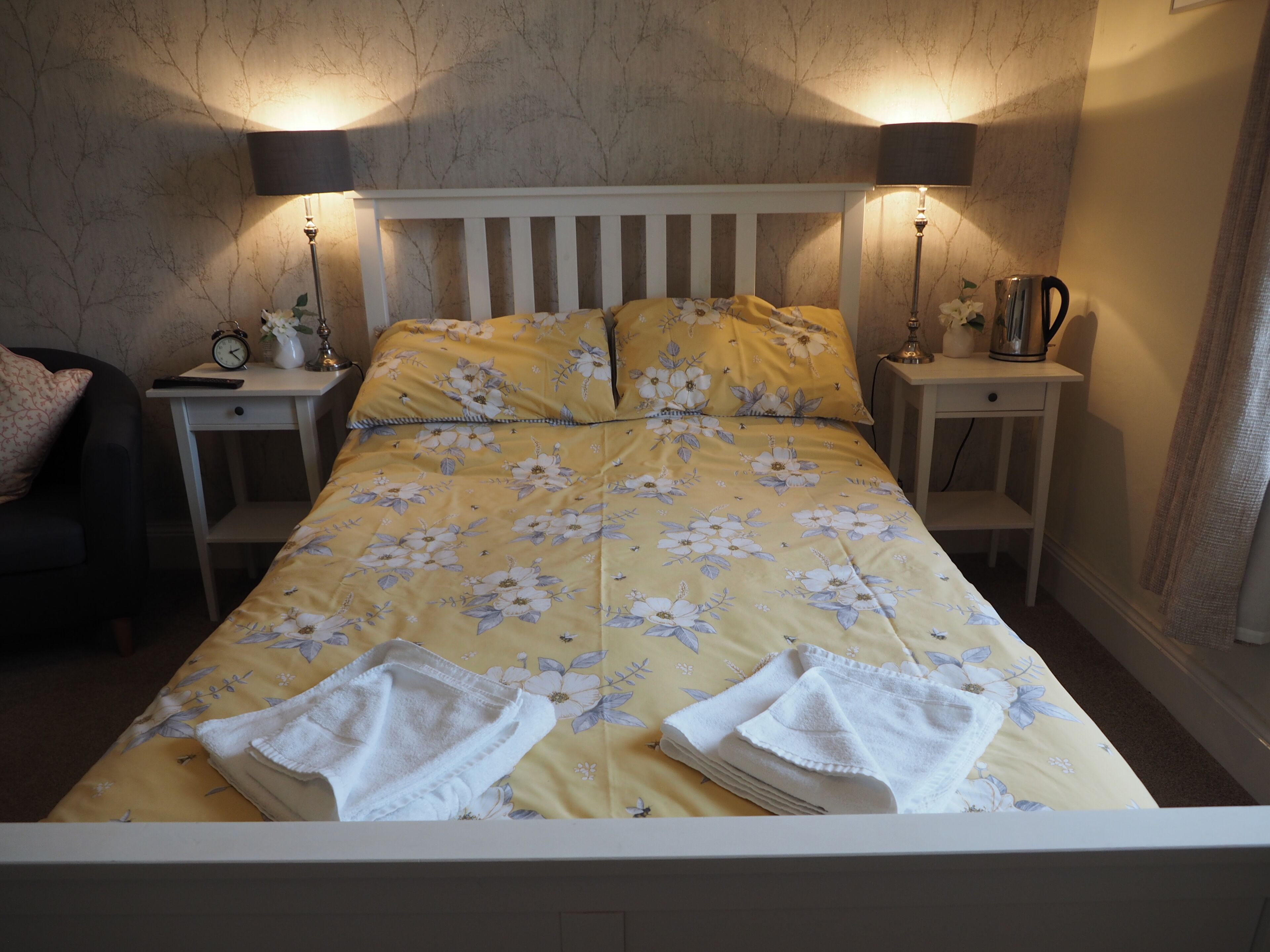 A neatly made bed with floral bedding, two bedside tables with lamps, a nightstand with a clock, and a chair with a cushion.