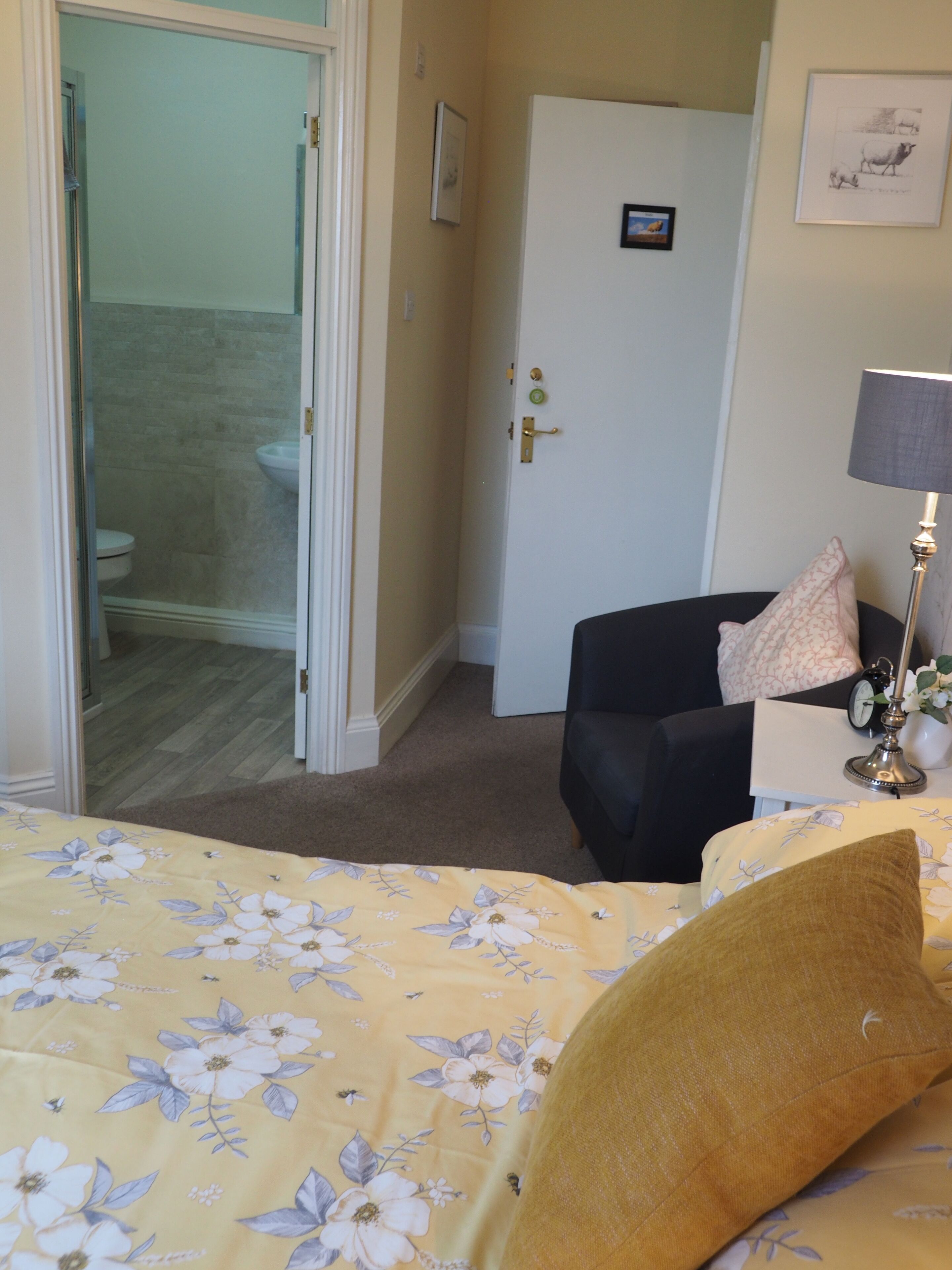 A hotel room with a bed, a chair, a desk with a lamp, a door, and a bathroom visible through an open door.