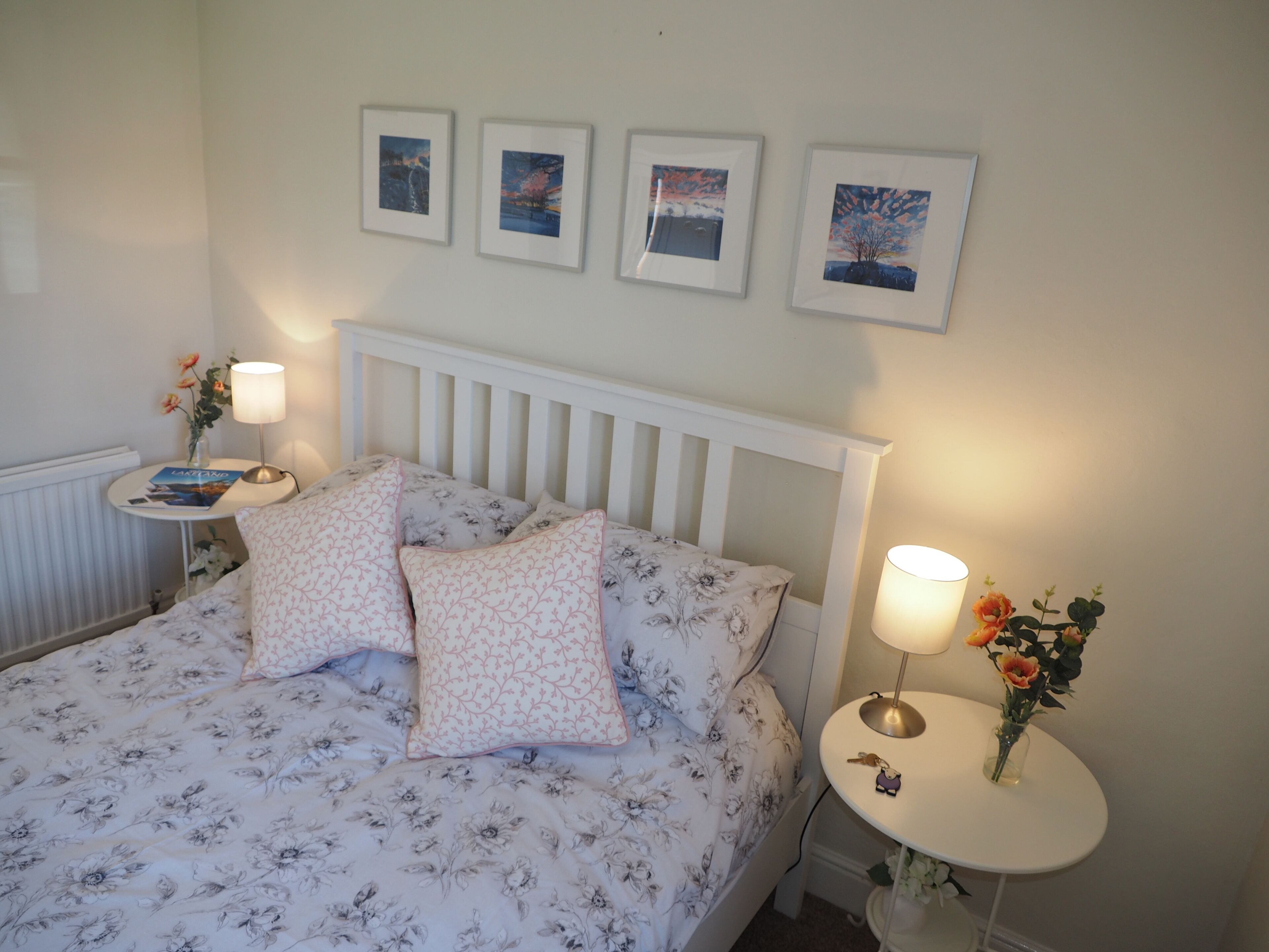 A neatly made bed with floral bedding and two pillows, a bedside table with a lamp and a book, and a small table with a vase of flowers.