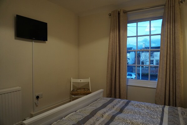 Un dormitorio con una cama, una silla, un televisor montado en la pared y una ventana con cortinas.