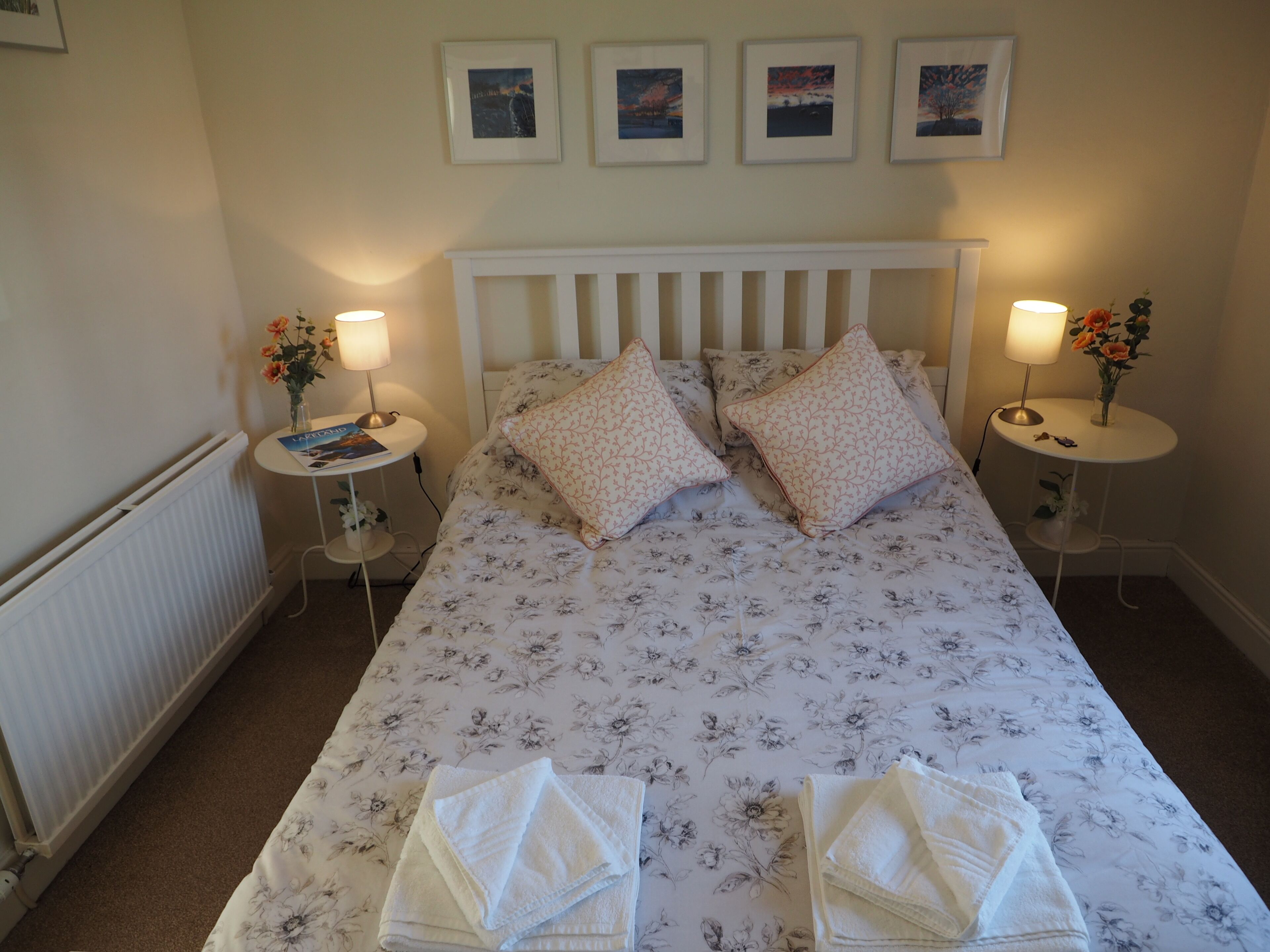Una cama bien hecha con ropa de cama floral, dos mesitas de noche con lámparas y flores, y cuadros en la pared.
