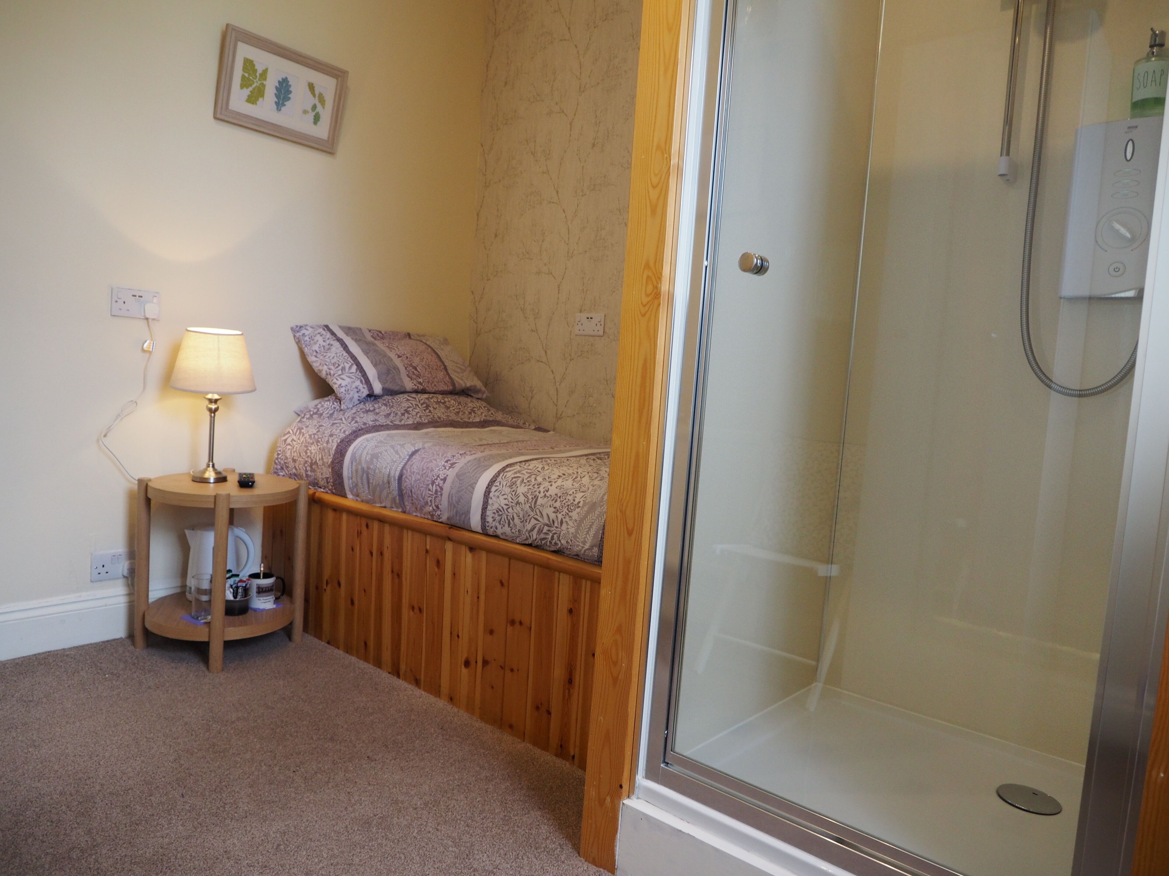 A bedroom with a bed, a bedside table with a lamp, a shower, and a wall-mounted power outlet.
