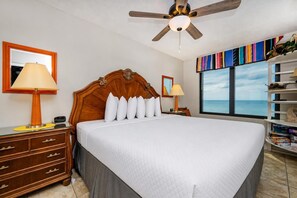 Quarto de hotel com uma cama grande, ventoinha no teto, vista para o oceano, estante e escrivaninha com luminária.