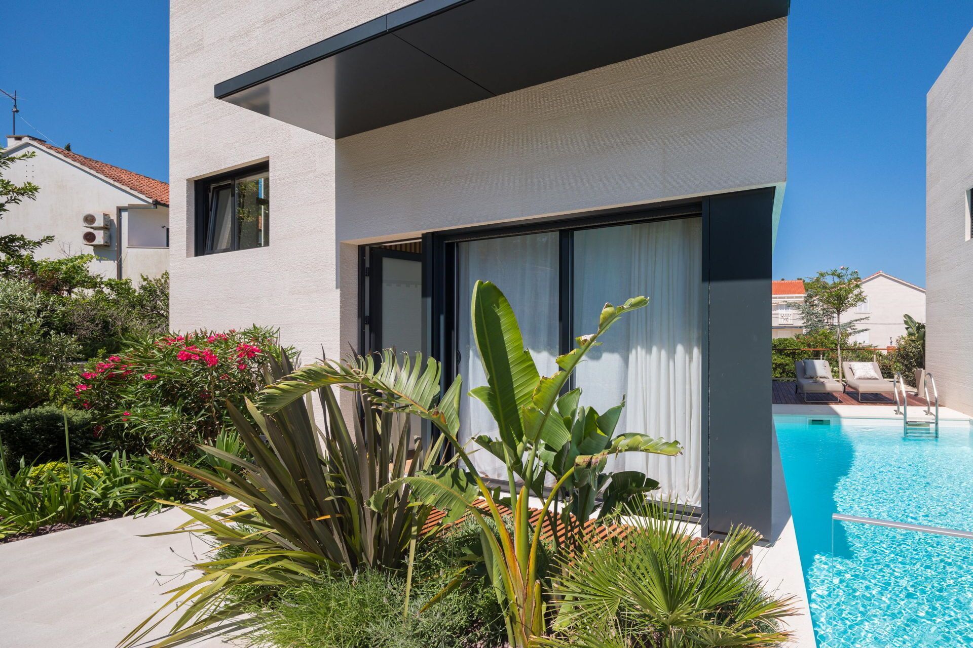 Un edificio moderno con piscina, rodeado de plantas tropicales y un cielo azul despejado.