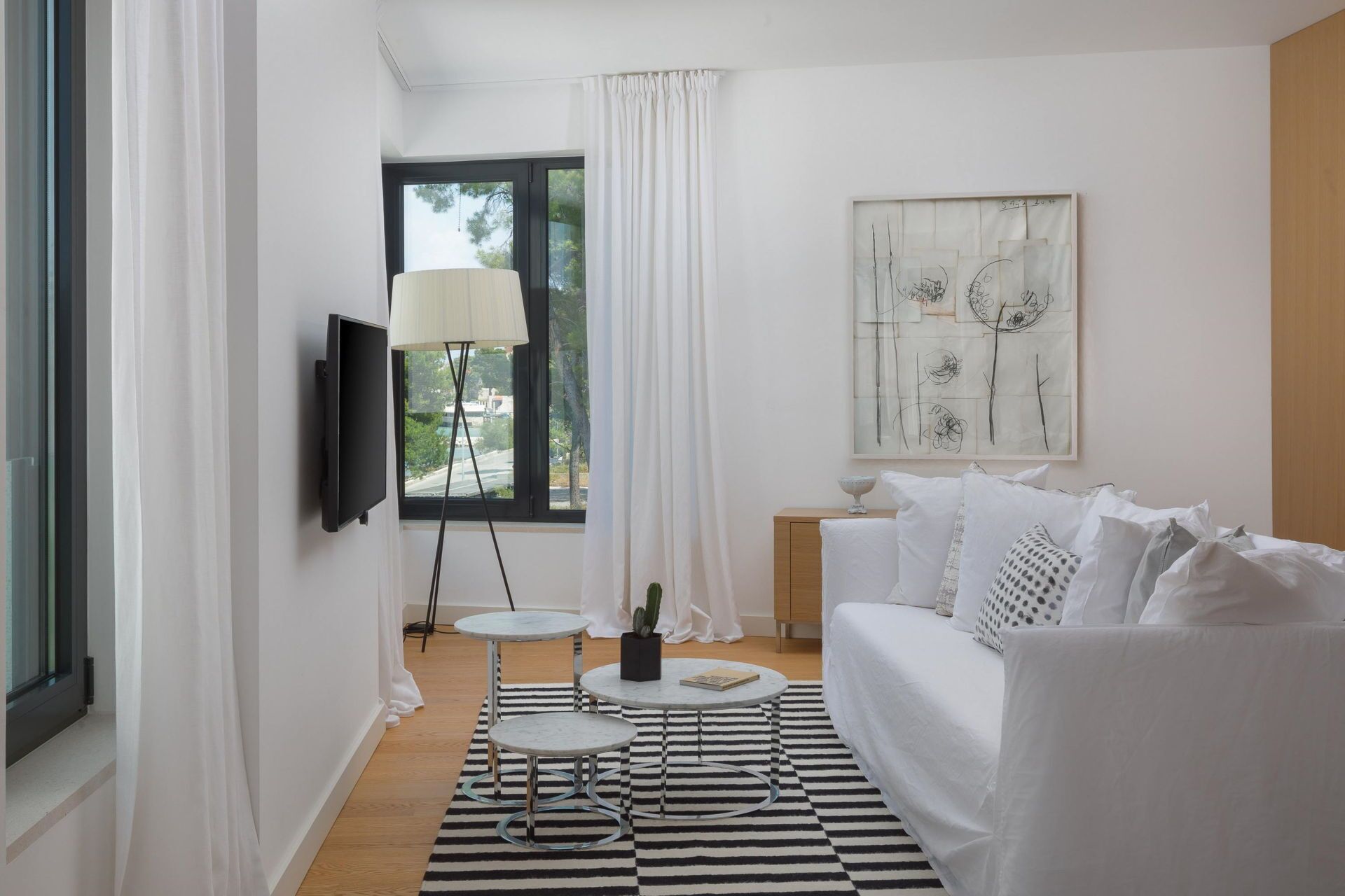 A modern living room with a white sofa, a small round table, a floor lamp, and a large window with curtains.