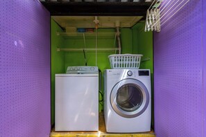 A compact laundry room with a washing machine and dryer, shelves above, and a laundry basket on the dryer. - Tropical Haven! 5BR w/ Lush Pool Backyard Retreat (Ponton)