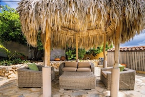 An outdoor seating area with wicker furniture under a thatched roof, surrounded by stone walls and greenery.