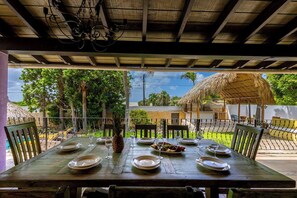 An outdoor dining area with a table set for two, surrounded by chairs, with a view of a thatched-roof structure and greenery in the background. - Tropical Haven! 5BR w/ Lush Pool Backyard Retreat (Ponton)