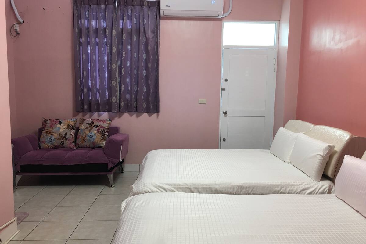 A bedroom with two beds, a purple sofa, a window with purple curtains, and an air conditioning unit.