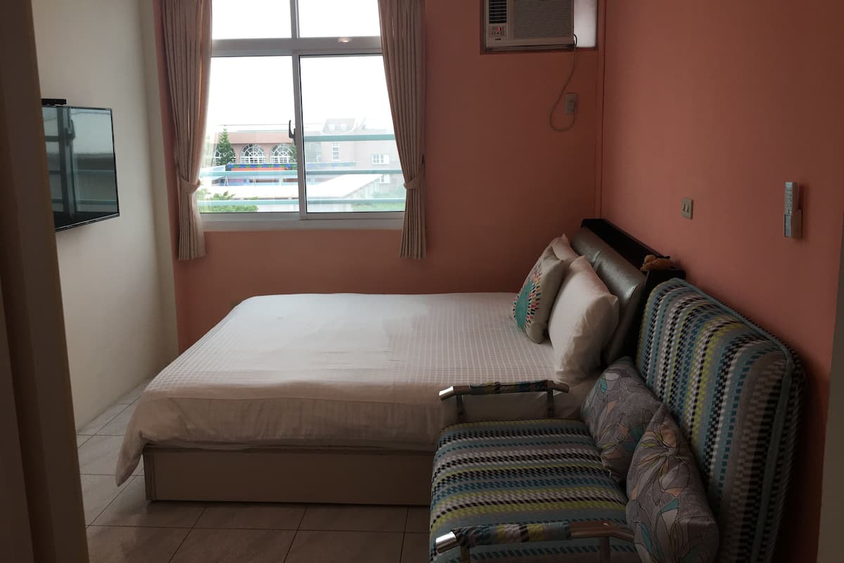 A bedroom with a bed, a sofa, a TV, an air conditioning unit, and a window with curtains.