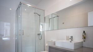 Un baño moderno con cabina de ducha de vidrio, inodoro blanco y un espejo grande sobre un lavabo blanco con dispensador de jabón.