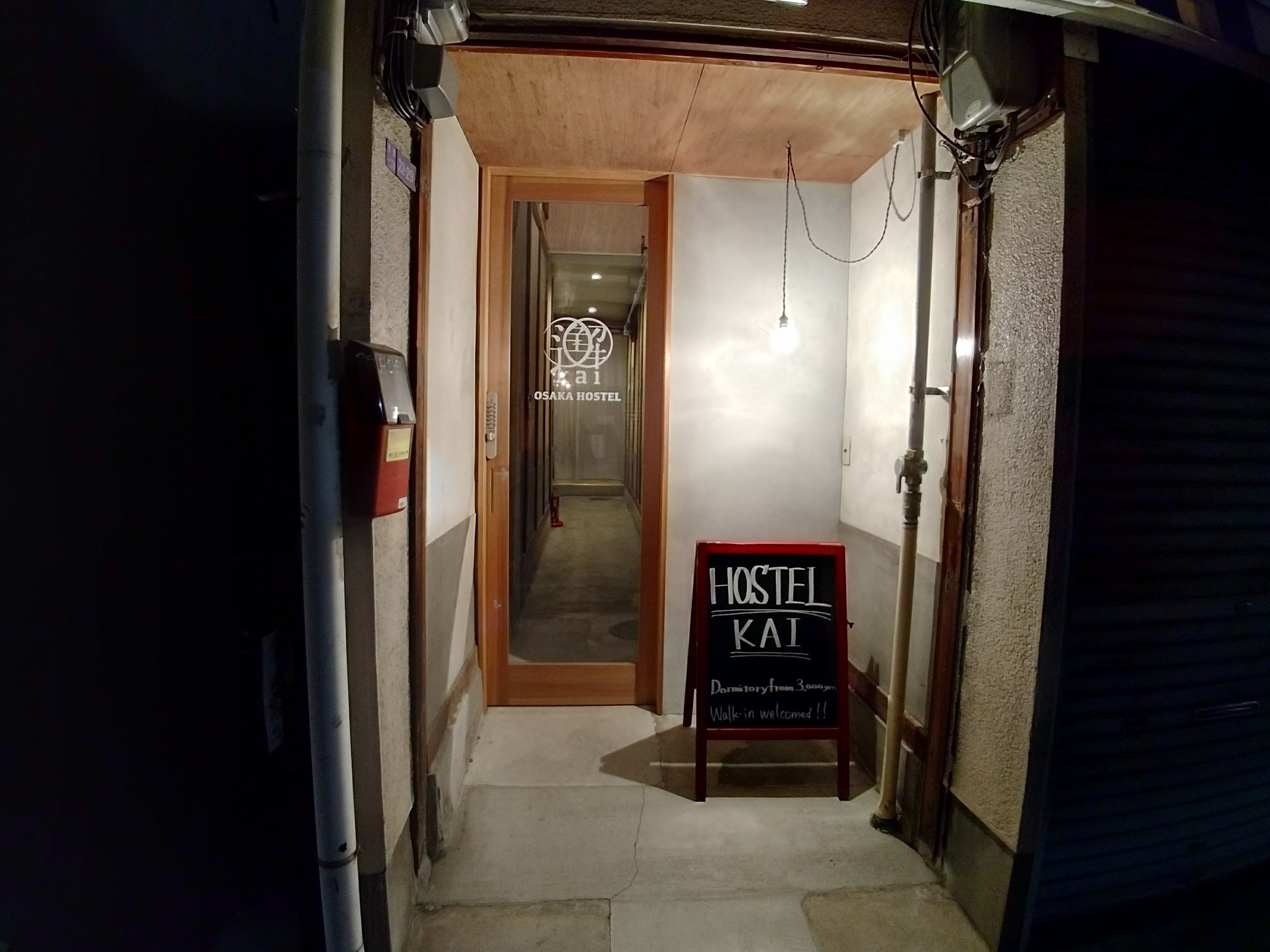 A narrow hallway leading to a room with a wooden door and a sign reading "OSAKA HOSTEL".