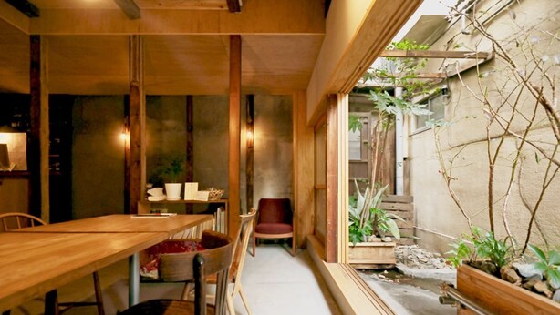Una mesa de comedor de madera con sillas, una silla roja y una mesita con cojín rojo. Se ve una ventana con plantas afuera.