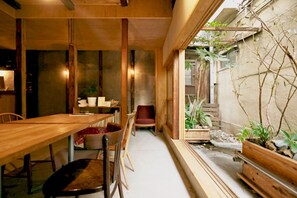 A wooden dining table with chairs, a red chair, and a small table with a red cushion. There is a window with plants outside. - OSAKA HOSTEL kai (Osaka)