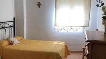 A room with two beds, a wooden dresser, a hanging lamp, a window with a curtain, and a wall-mounted cross.