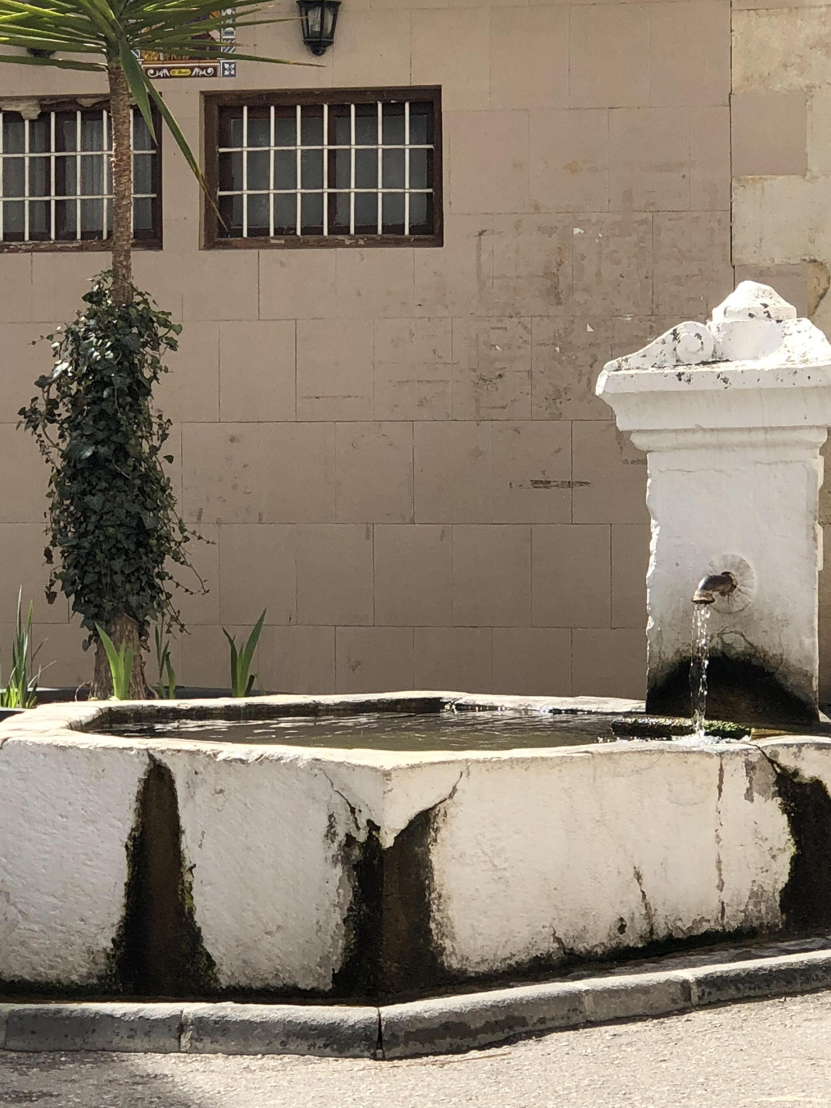 Une fontaine d’où jaillit de l’eau, entourée d’une bordure en béton, se dresse devant un bâtiment dont la façade est recouverte de carreaux et dont les fenêtres sont grillagées.