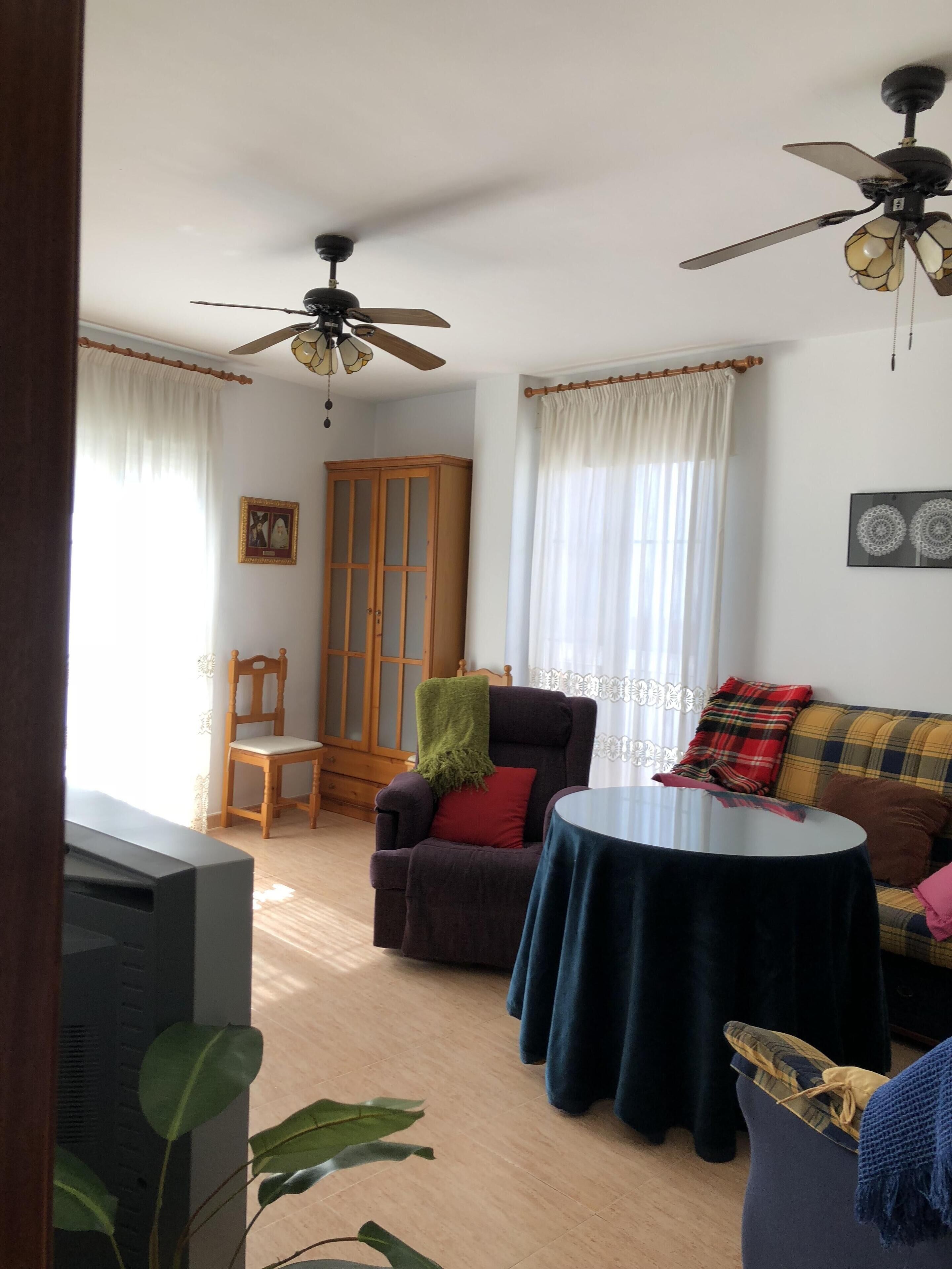 Ein Wohnzimmer mit Sofa, Sessel und Tisch. Im Raum befindet sich ein Holzschrank und ein Fernseher.