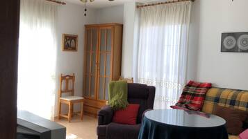 Ein Wohnzimmer mit Sofa, Sessel und Tisch. Im Raum befindet sich ein Holzschrank und ein Fernseher.