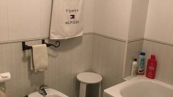 Une salle de bain équipée d’un bidet, de toilettes et d’une baignoire. Des serviettes sont accrochées à des crochets et des articles de toilette sont disposés sur une étagère.