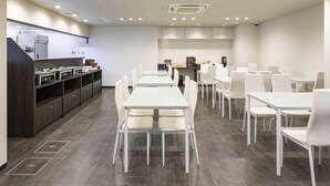 A modern dining area with white tables and chairs, a kitchen in the background, and a dark wooden counter with various items. - Hotel Axia Inn Osaka Uehonmachi (Osaka)